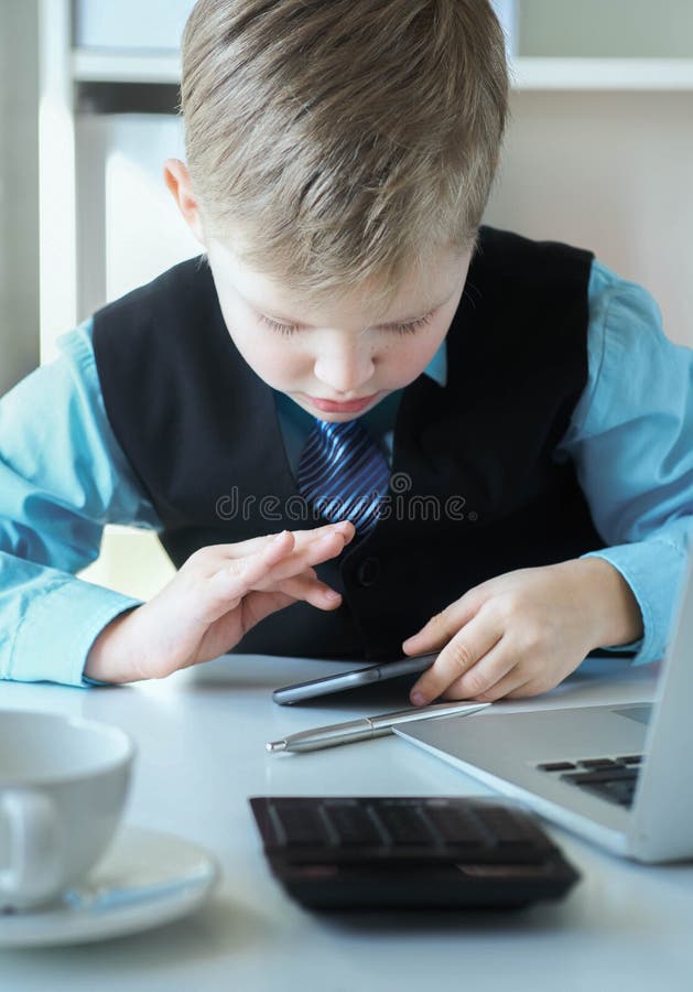 Smart liten pojke i kostym som arbetar med den bärbara datorn och som rör på telefonskärmen känslomässigt royaltyfria bilder