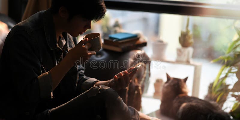 A smart man drinking hot coffee while using a smartphone in hand and sitting on leather couch next to his lovely cat.