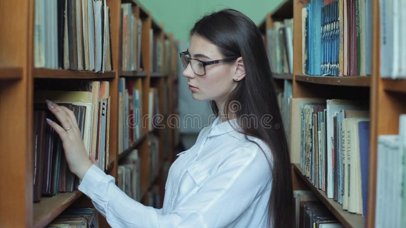 Smart se och härlig ung flicka i den vita skjortan och exponeringsglas i collageskolaarkivet som väljer boken att läsa arkivfilmer