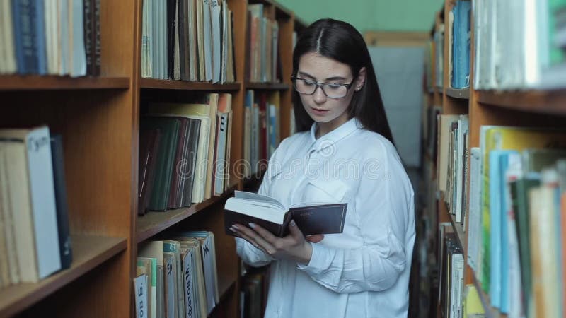 Smart se och härlig ung flicka i den vita skjortan och exponeringsglas i collageskolaarkivet som väljer boken att läsa stock video
