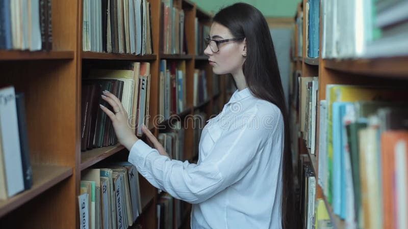 Smart se och härlig ung flicka i den vita skjortan och exponeringsglas i collageskolaarkivet som väljer boken att läsa stock video