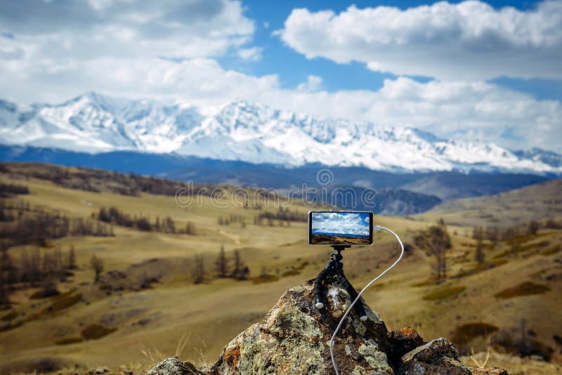 Smartphone on small tripod takes pictures of the mountain range and the clouds above the snowy peaks.  Blurred background, focus
