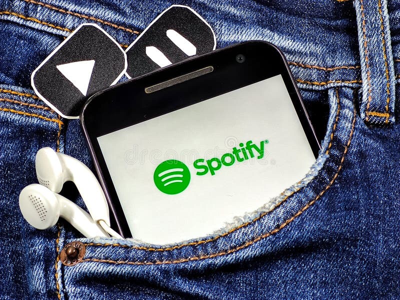 Smartphone with Spotify app on the screen in jeans pocket with pause and play buttons and headphones