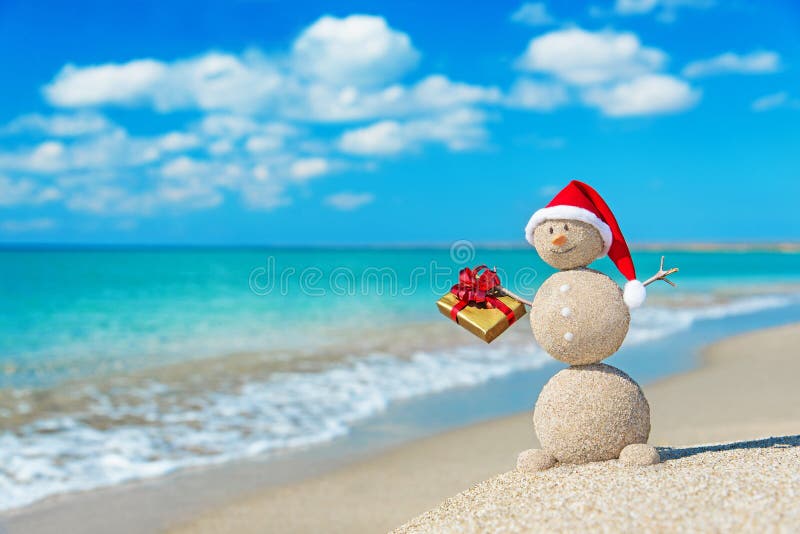 Smiley zandige sneeuwman bij strand in Kerstmishoed met gouden gift