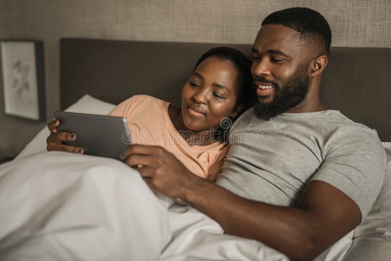 Smiling young African American couple watching videos on a tablet together while lying in their pajamas in their bed in the morning. Smiling young African American couple watching videos on a tablet together while lying in their pajamas in their bed in the morning