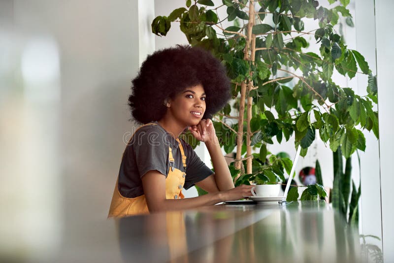 Smiling African lady looking through window sitting at cafe table using laptop.