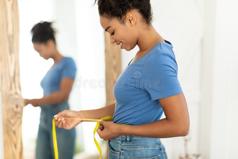 Smiling African Woman Measuring Waist After Slimming Standing At Home. Smiling African Woman Measuring Waist With Tape After Slimming And Successful Weight Loss