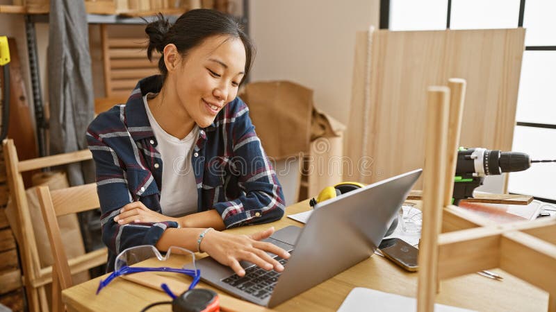 Smiling asian woman using laptop in a woodworking workshop surrounded by tools and timber royalty free stock photos