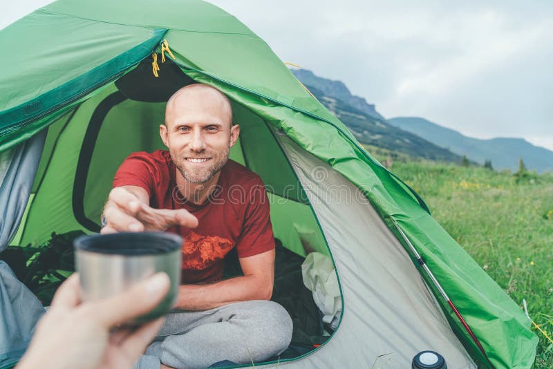 Smiling bald Man in the green tent taking the thermos teacup for morning tea-drinking with mountain background. Active people