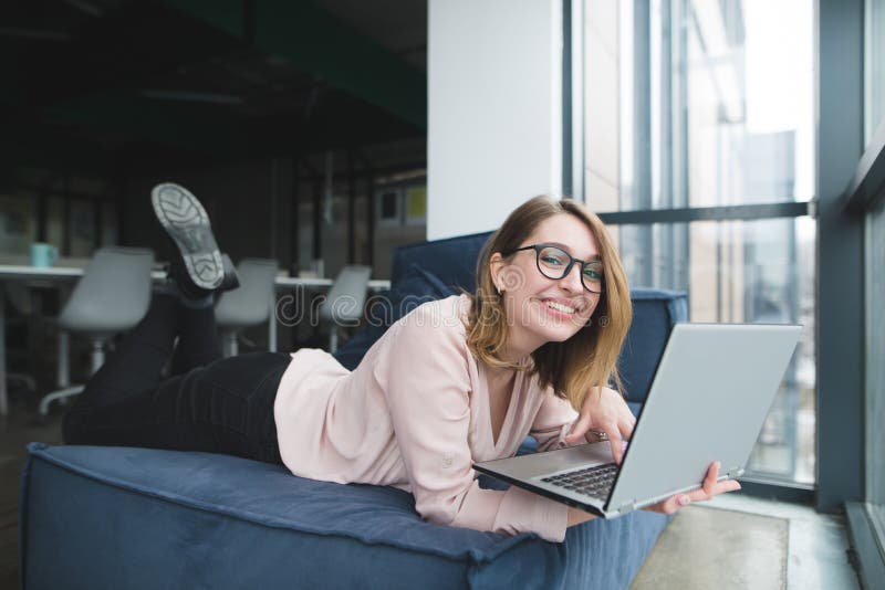 Smiling beautiful girl lying on a sofa with a laptop in her hands at the background of office dressing