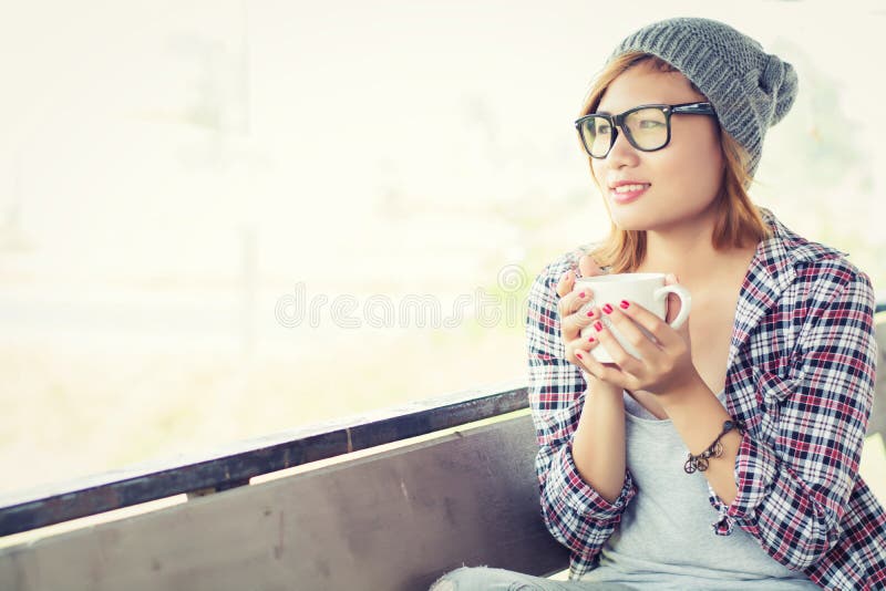 Smiling beautiful hipster woman sitting on bench with morning co