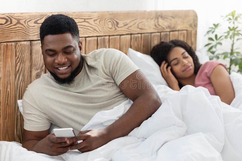 Smiling black men chatting with lover on smartphone while his wife sleeping by him in bed. African american husband having affair, sending messages to another woman. Cheating in marriage concept. Smiling black men chatting with lover on smartphone while his wife sleeping by him in bed. African american husband having affair, sending messages to another woman. Cheating in marriage concept