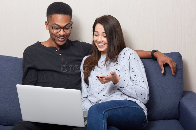 Smiling black young man and dark haired woman relax on couch, watching movie on laptop, happy multicultural couple resting on sofa