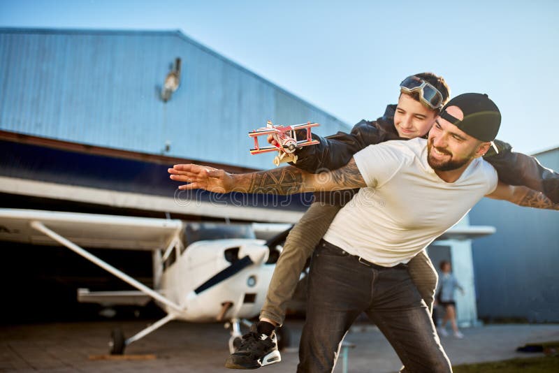 Smiling boy with model plane, riding on father`s back, playing outside hangar