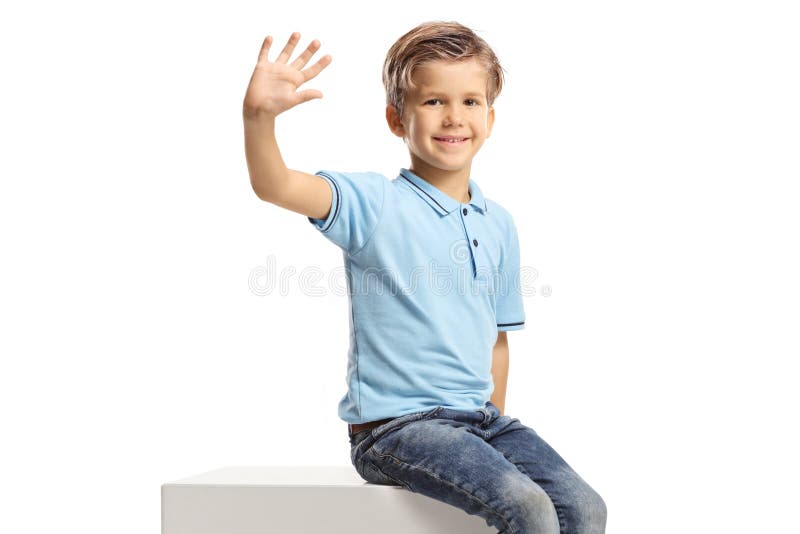Smiling boy sitting on a white cube and waving isolated on white background