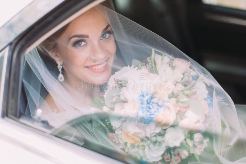 Smiling bride under the veil is holding the wed bouquet while sitting in the car.