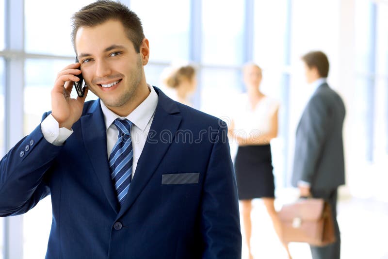 Smiling businessman in office with colleagues in the background and using mobile royalty free stock photography Smiling businessman in office with colleagues in the background and using mobile royalty free stock photography