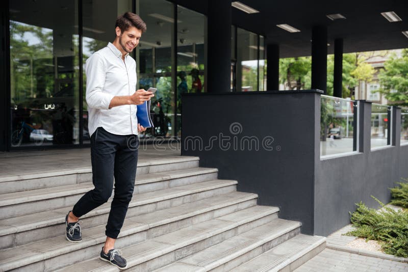 Smiling attractive young businessman walking and using mobile phone near business center. Smiling attractive young businessman walking and using mobile phone near business center