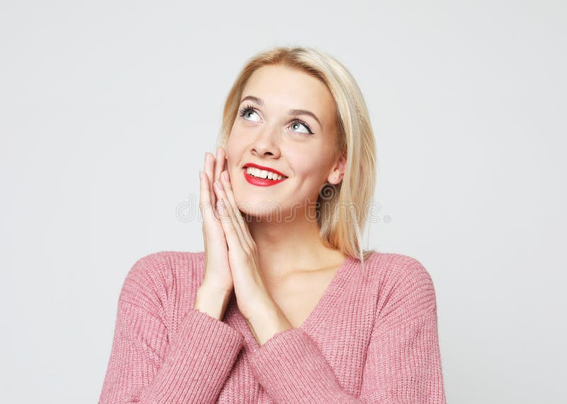 Smiling blond woman wearing pink sweater touching perfect skin and smiling, standing on grey background.