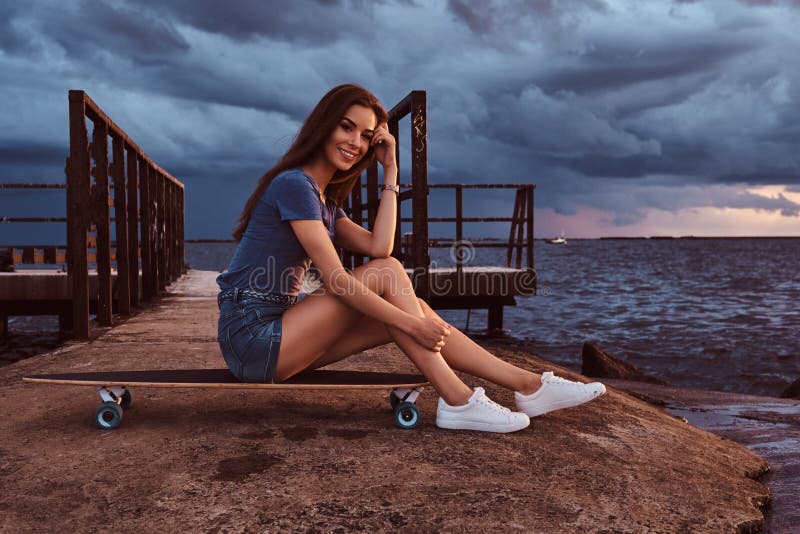 Sensual Girl Sitting On A Skateboard On The Beach Is Enjoying Amazing Dark Cloudy Weather During