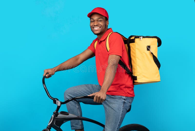 Smiling delivery delivery man riding bicycle at studio. Delivering Orders On Time. Portrait of cheerful black male courier in red cap and uniform riding bicycle