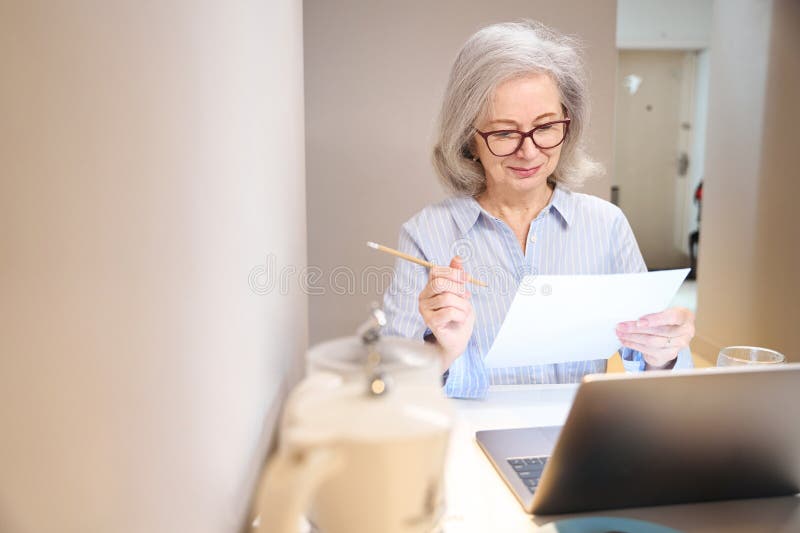 Smiling Elderly Lady Works with Documents and Laptop Kitchen Table