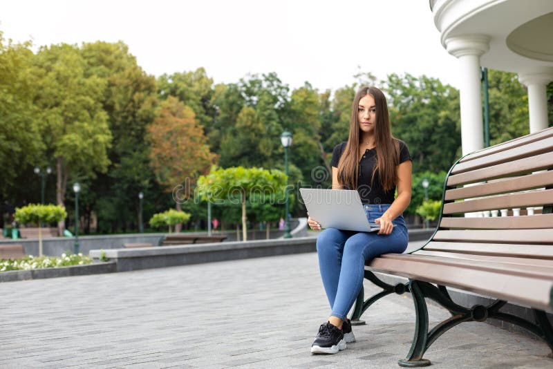 Education, business, technology, and Internet concept — Smiling European dark-haired girl in a black T-shirt and blue jeans sitting on a bench in the park in the summer and holding a laptop computer