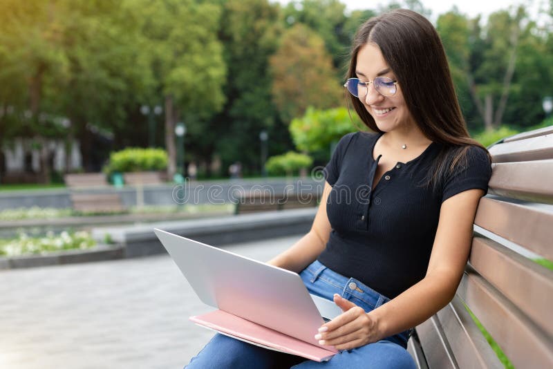 Education, technology, and Internet concept — A smiling European dark-haired student girl in modern clothes, sitting on a park bench in the summer and looking at her laptop with notebooks