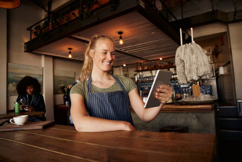 Smiling female waiter standing leaning on table using digital tablet in modern cafe royalty free stock photography