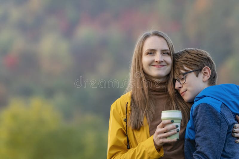 Smiling girl drinks coffee or tea and hugs son. Loving mother and child spend time together walking autumn nature