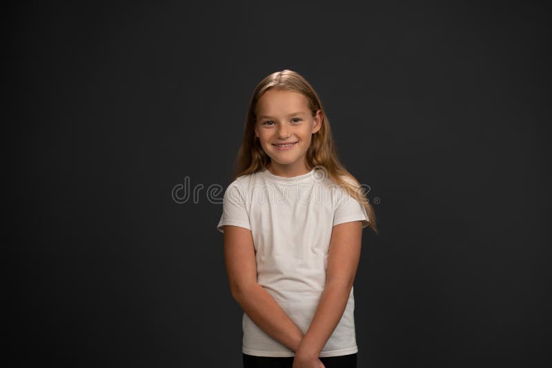 Smiling girl looking timid or frustrated at the camera wearing white t-shirt isolated on dark grey or black background