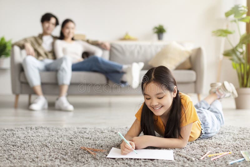 Smiling japanese youngsters girl study at home, writing, drawing in notebook and millennial parents sits and rest