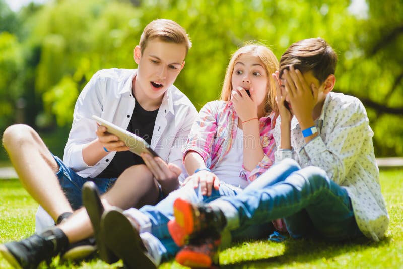 Smiling kids having fun and look to tablet at grass. Children playing outdoors in summer. teenagers communicate outdoor