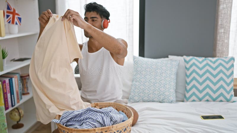A smiling latino man in a tank top enjoys music on red headphones while folding laundry in his well-lit bedroom stock video