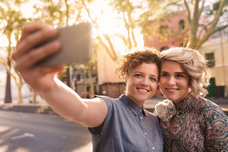 Smiling Lesbian Couple Taking Selfies Together On A City Sidewalk Stock