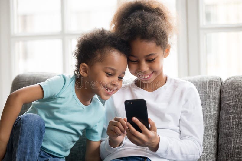 Happy little mixed race kids sit on couch play game on smartphone together, smiling small black brother and sister have fun hold using cellphone, watch funny cartoon. Children and technology concept. Happy little mixed race kids sit on couch play game on smartphone together, smiling small black brother and sister have fun hold using cellphone, watch funny cartoon. Children and technology concept