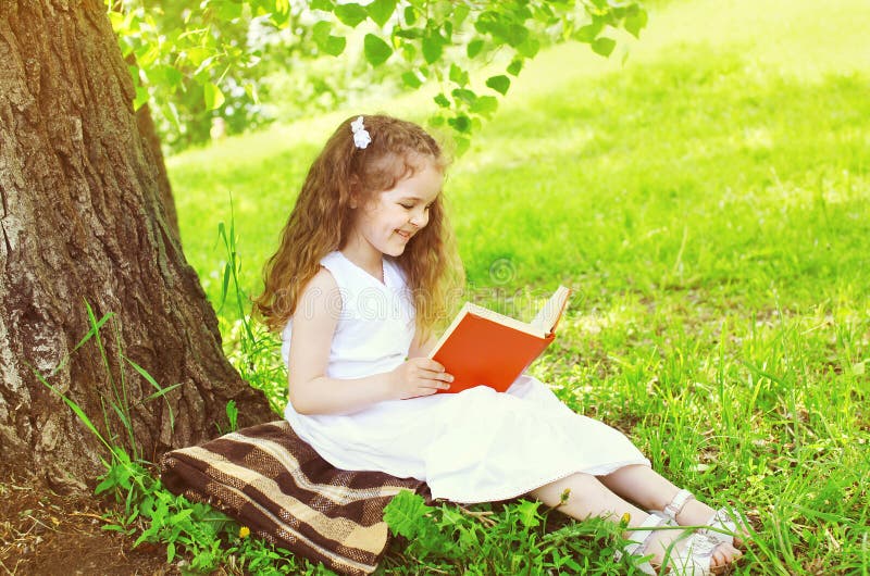 Smiling little girl child reading a book on the grass near tree