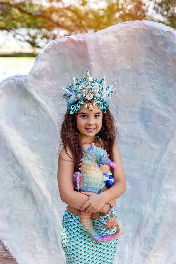 A smiling little girl in a turquoise mermaid costume and a crown is standing outdoors, in a large seashell, holding a colorful seahorse toy. Vertical. Close up. Copy space. A smiling little girl in a turquoise mermaid costume and a crown is standing outdoors, in a large seashell, holding a colorful seahorse toy. Vertical. Close up. Copy space