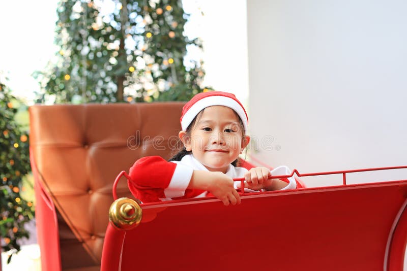 Smiling little kid girl in santa costume dress sitting on red sledge christmas background. Merry Christmas and happy new year royalty free stock photo Smiling little kid girl in santa costume dress sitting on red sledge christmas background. Merry Christmas and happy new year royalty free stock photo