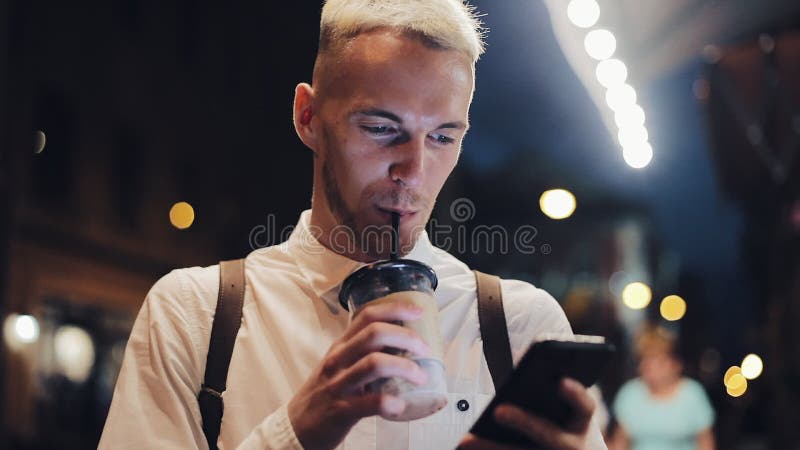 Smiling man usingn smartphone in night city. Handsome young man using smartphone at night and drinking ice coffee stock video footage