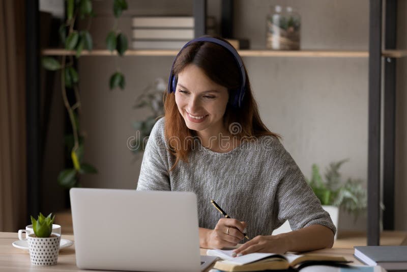 Smiling millennial Caucasian girl in headphones sit at desk look at laptop screen study online, happy smart young woman in earphones take web course or training on computer, distant education concept. Smiling millennial Caucasian girl in headphones sit at desk look at laptop screen study online, happy smart young woman in earphones take web course or training on computer, distant education concept