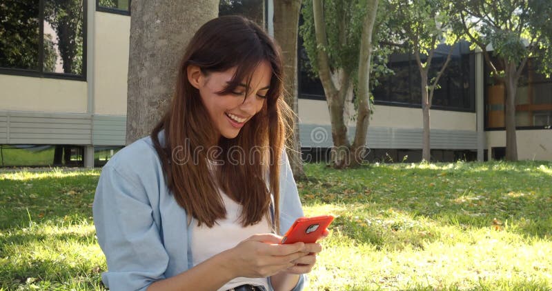 Smiling millennial young woman chats with a smart phone sitting in a park