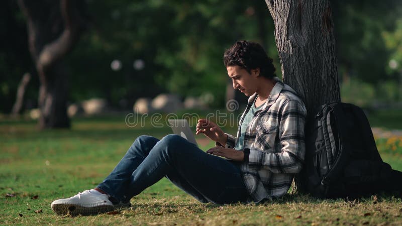 Smiling mixed race casual student guy e learning remotely use laptop outdoor at summer sunny park stock footage