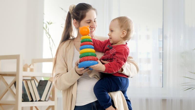 SMiling mother holding her baby som and giving him colorful toy tower or piramid