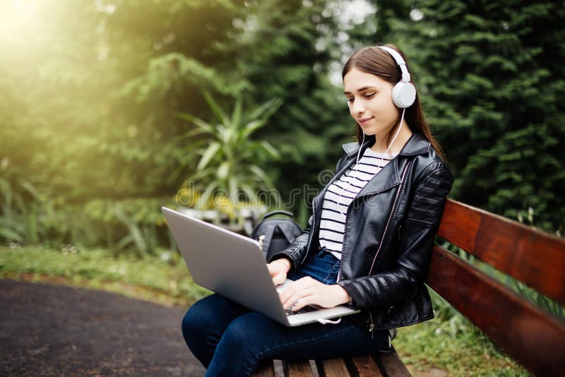 Smiling student sitting on bench listening music and holding laptop in park at school. Smiling student sitting on bench listening music and holding laptop in park at school