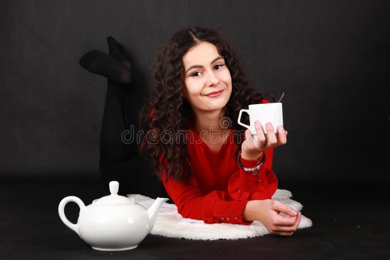 Smiling teenage girl in red blouse and black pantyhose drinking tea from white cap while lying on white sheep fur. Teapot aside of her. Black background. Tea party concept. Smiling teenage girl in red blouse and black pantyhose drinking tea from white cap while lying on white sheep fur. Teapot aside of her. Black background. Tea party concept.