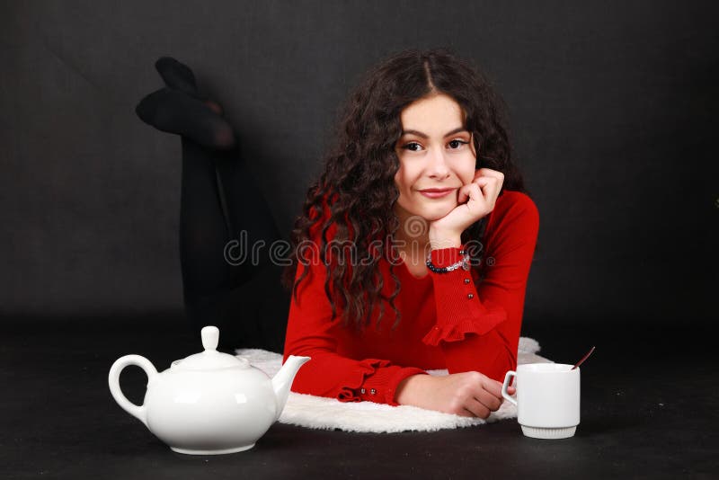 Smiling teenage girl in red blouse and black pantyhose drinking tea from white cap while lying on white sheep fur. Teapot aside of her. Black background. Tea party concept. Smiling teenage girl in red blouse and black pantyhose drinking tea from white cap while lying on white sheep fur. Teapot aside of her. Black background. Tea party concept.