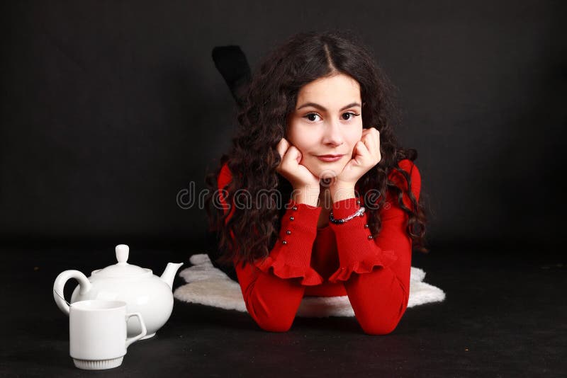 Smiling teenage girl in red blouse and black pantyhose drinking tea from white cup while lying on white sheep fur. Teapot aside of her. Black background. Tea party concept. Smiling teenage girl in red blouse and black pantyhose drinking tea from white cup while lying on white sheep fur. Teapot aside of her. Black background. Tea party concept.