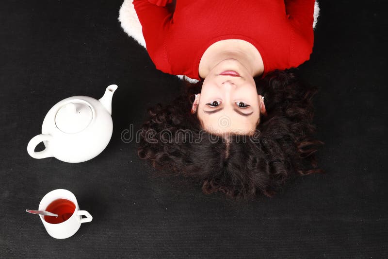 Smiling teenage girl in red blouse and black pantyhose  lying aside tea cup and teapot. Black background. Tea party concept. Smiling teenage girl in red blouse and black pantyhose  lying aside tea cup and teapot. Black background. Tea party concept.