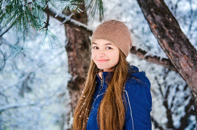 Smiling teenager girl in a blue down jacket and beige hat on a background of snowy fir branches. Winter, Christmas, New Year